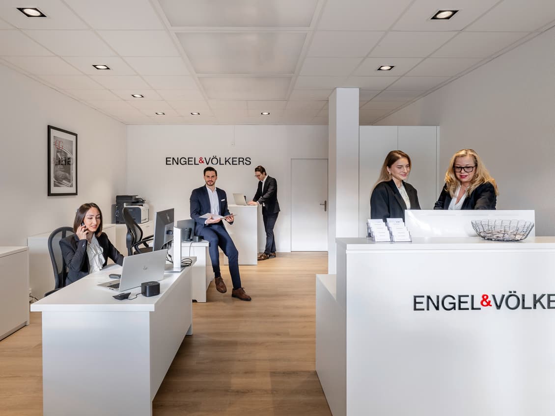 Engel & Völkers, extérieur d'une agence immobilière. Une chaise rouge avec le logo de l'entreprise est placée devant l'entrée. Des annonces immobilières sont affichées en vitrine.